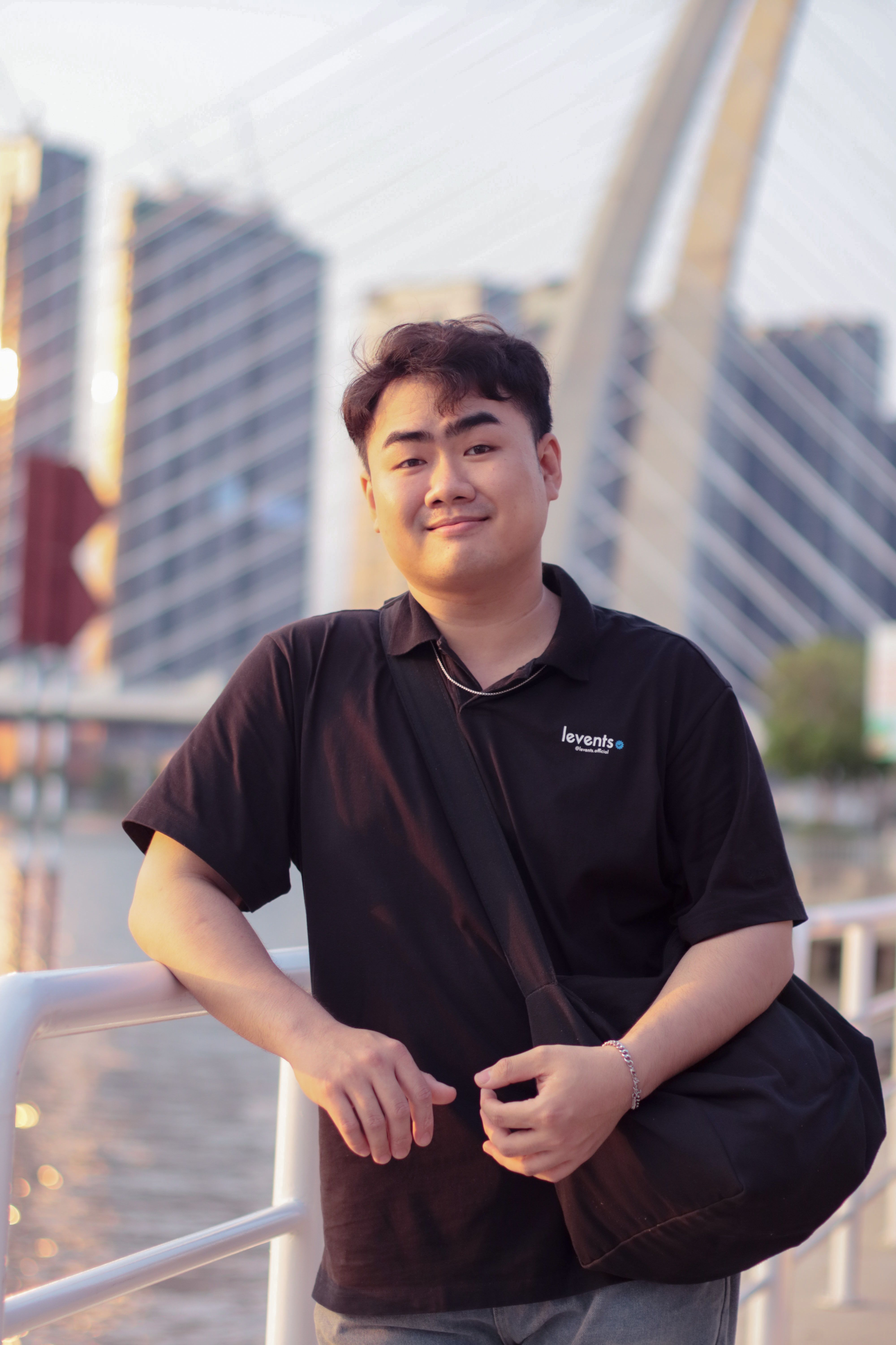 A cheerful young man smiling on a bridge with a cityscape backdrop, captured at sunset.