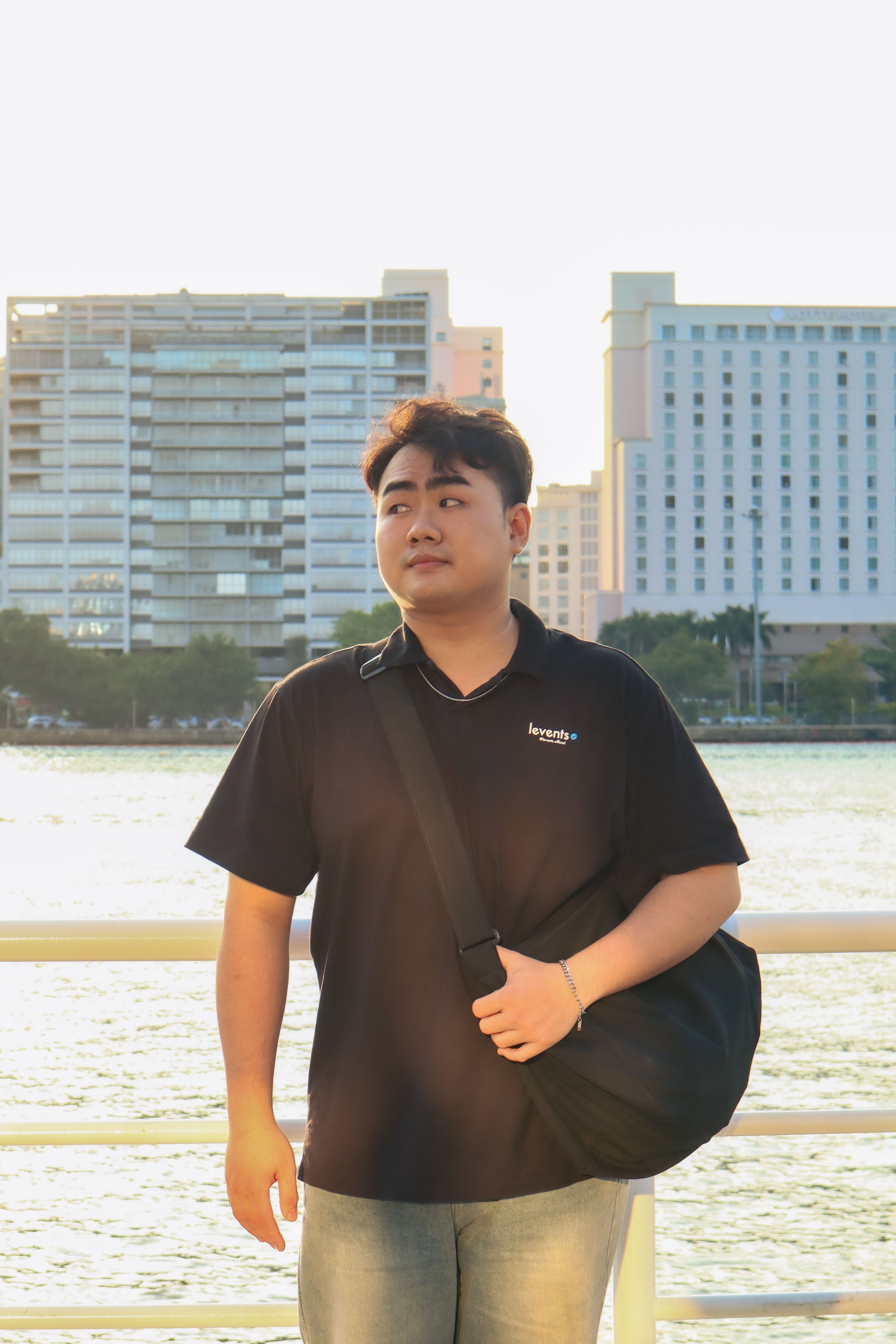 A young adult male standing by a waterfront with city buildings in the background during the day.