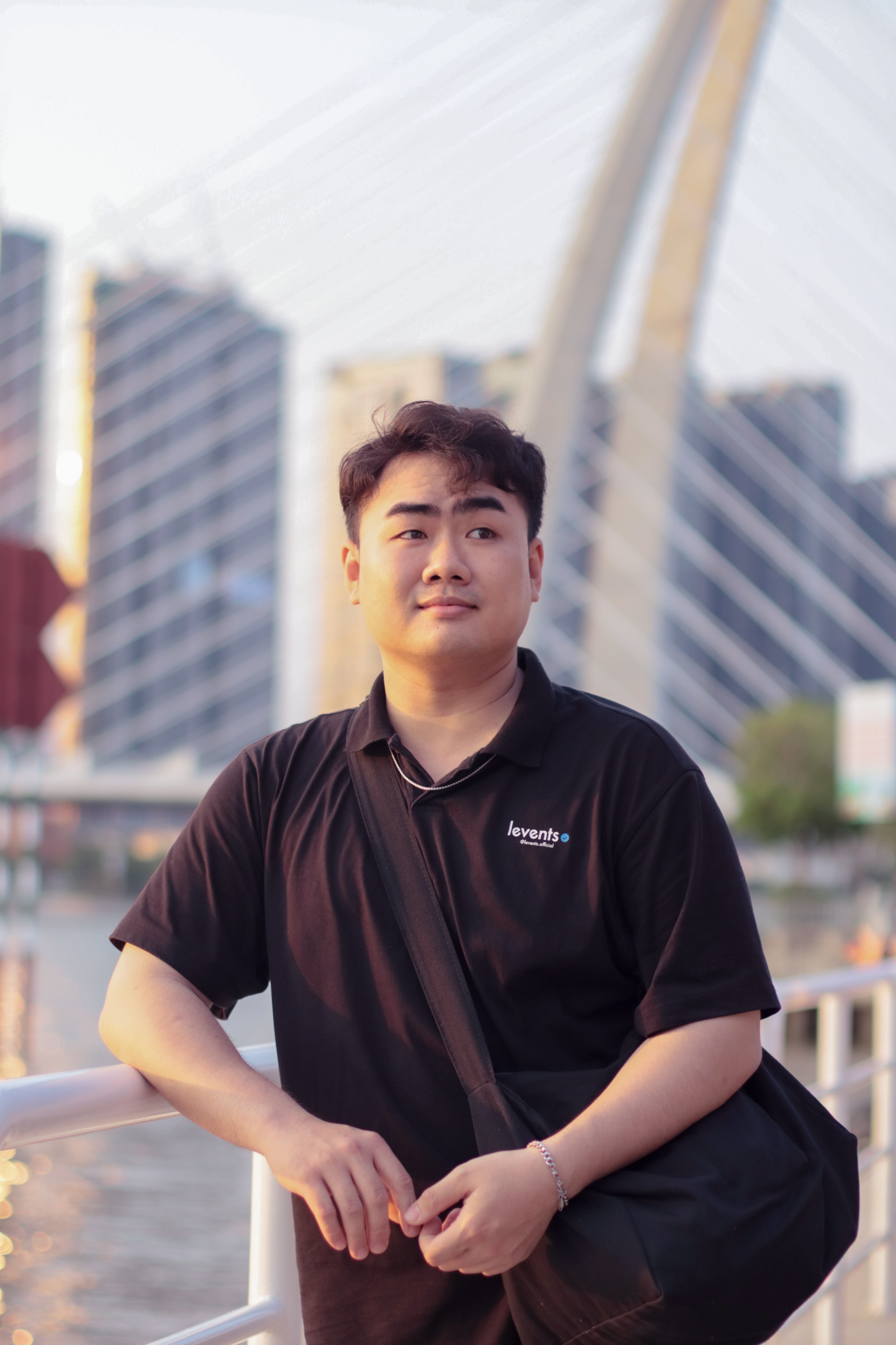 A young man in a black shirt stands on a bridge with a cityscape backdrop during the day.