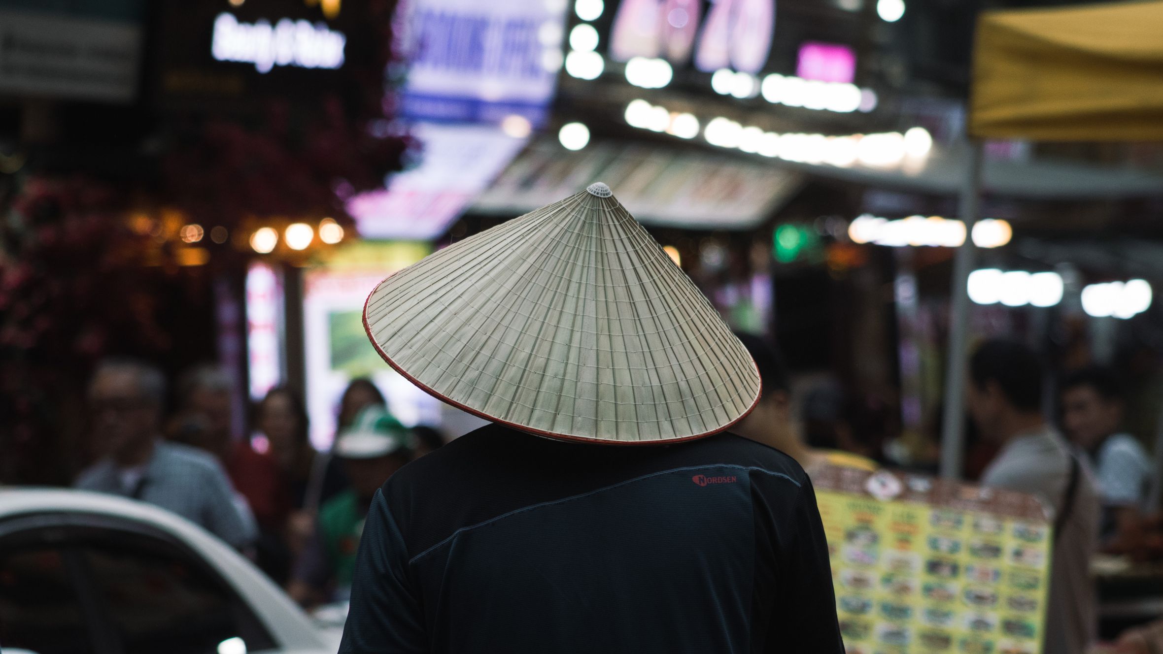 Man wearing traditional Vietnamese hat at Hanoi night market. Vibrant street life captured.