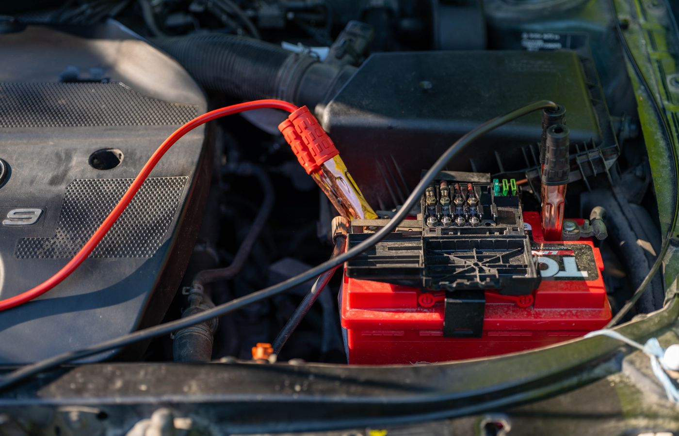 Close-up of a car battery with attached jumper cables in an engine bay.