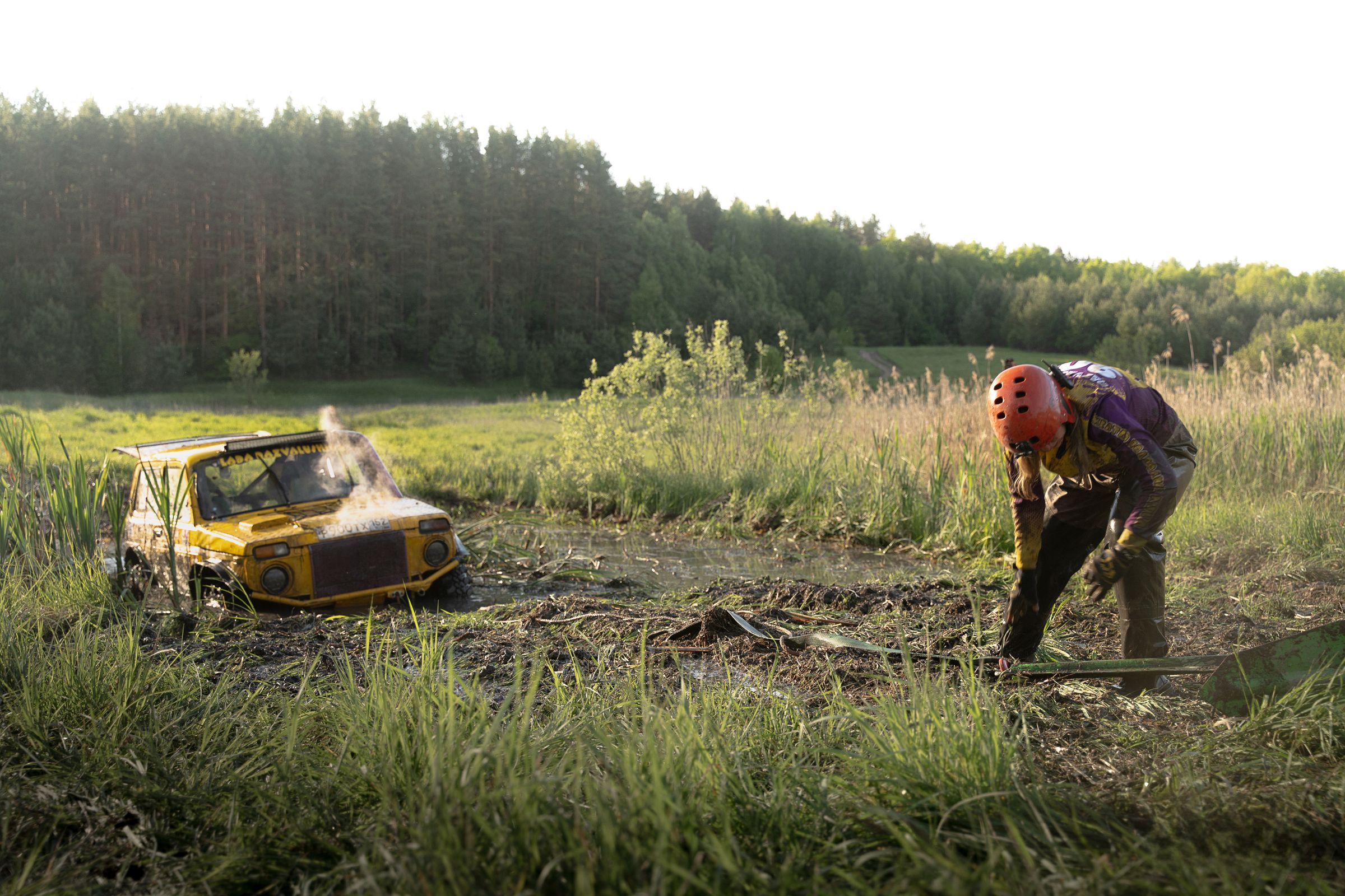 Off-road 4x4 stuck in a muddy bog at golden hour with a driver setting up a recovery strap