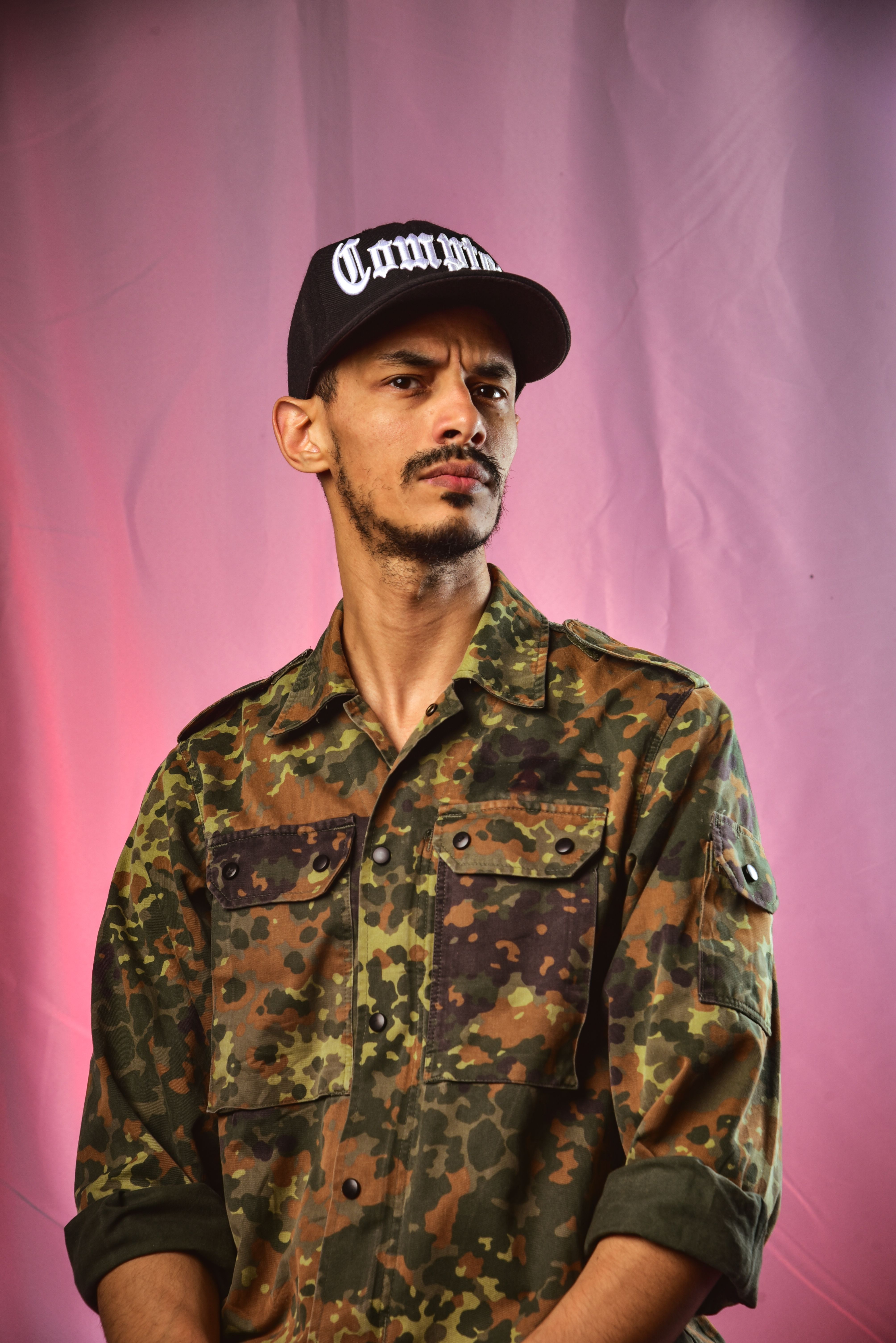 Man wearing camouflage shirt and cap with 'Common' text, set against pink background.