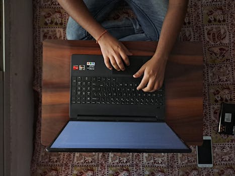 A person sits on the floor, typing on a laptop placed on a wooden table.