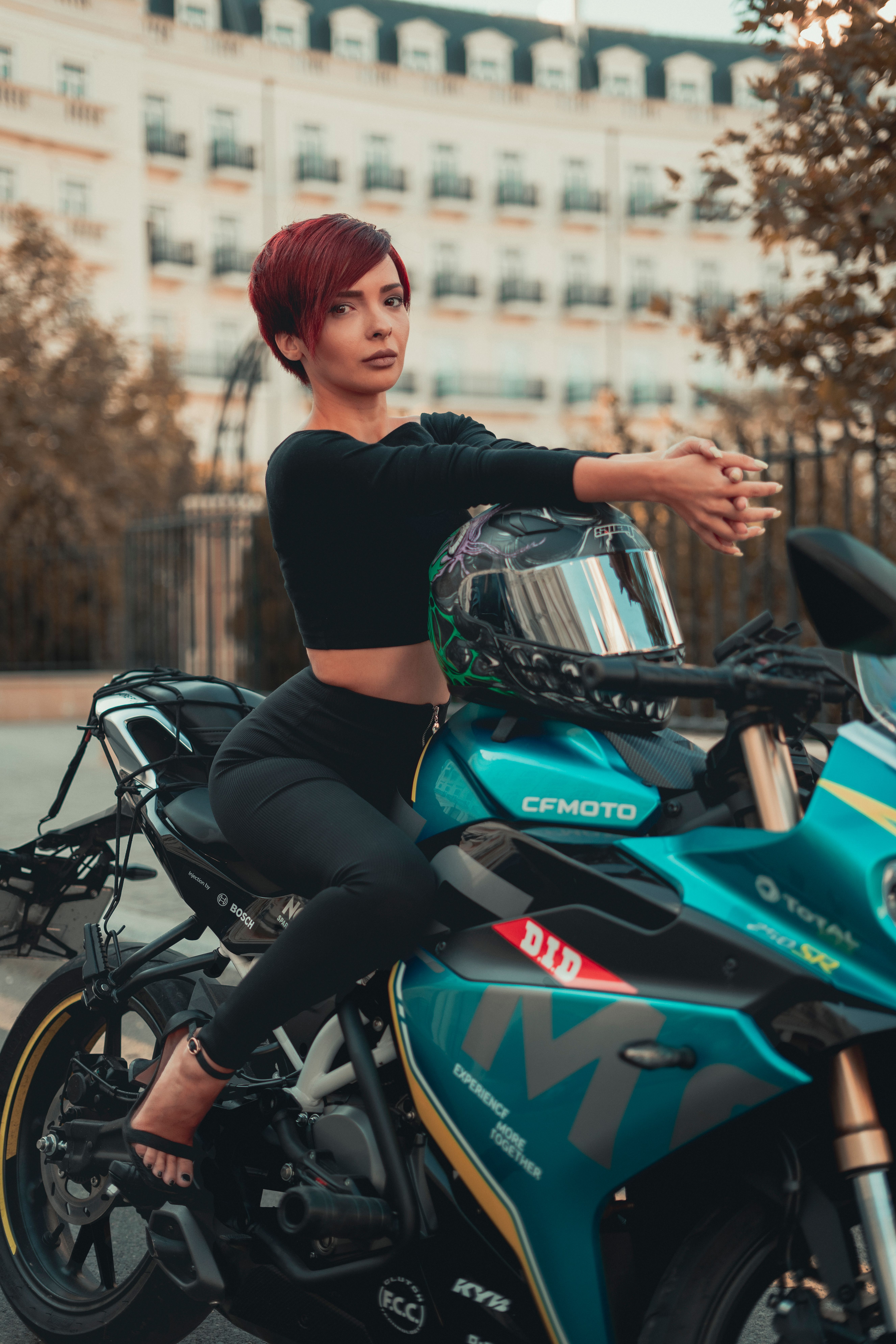 Stylish woman with short hair poses confidently on a modern motorcycle in an urban backdrop.