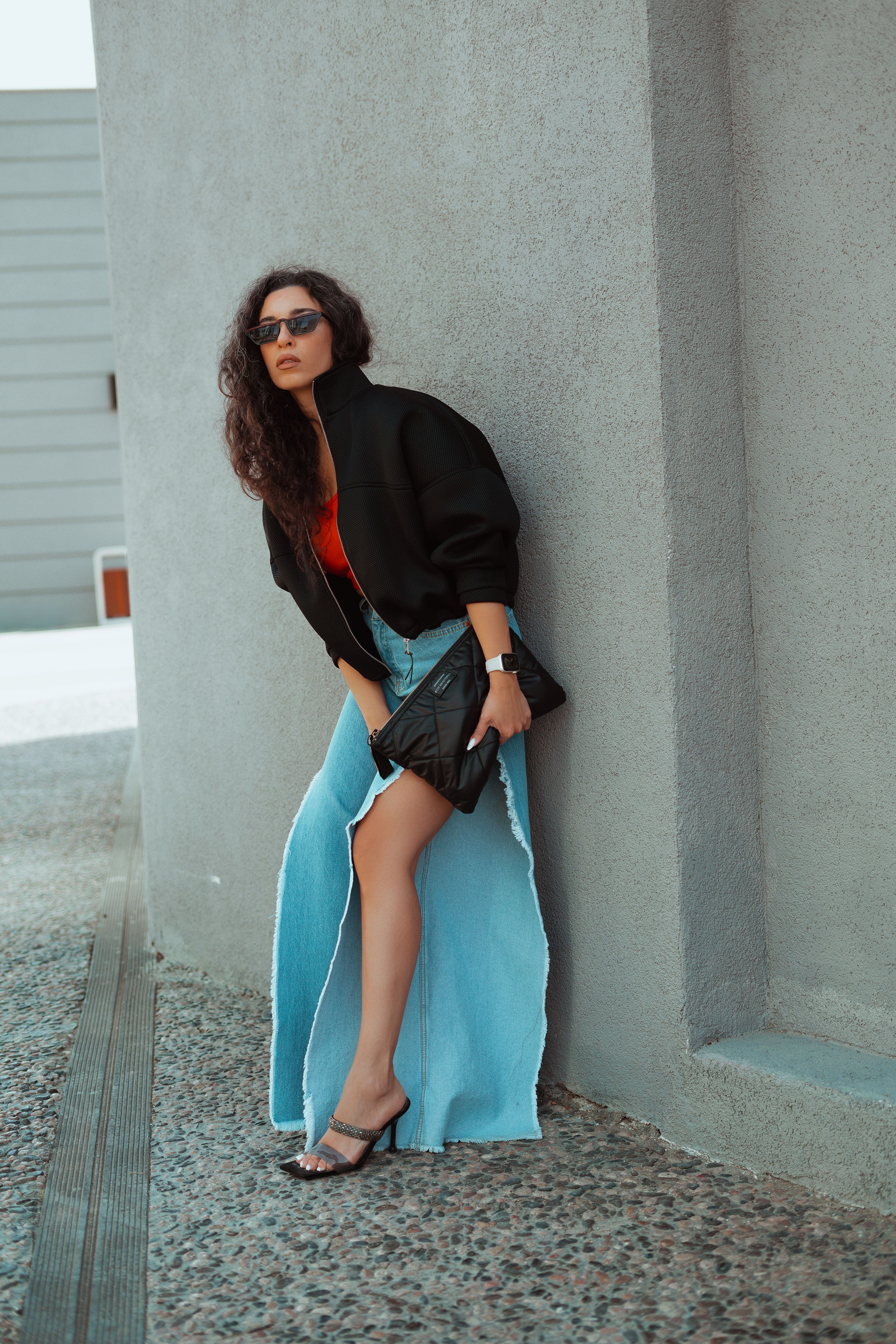 Mulher elegante com saia jeans e jaqueta posando perto de uma parede ao ar livre.