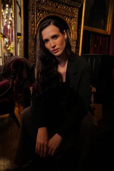 Sophisticated woman seated in an ornate, dimly lit room with rich decor.