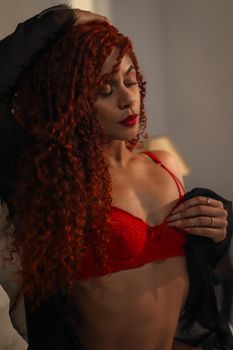 Elegant woman with curly red hair in red lingerie poses confidently indoors.
