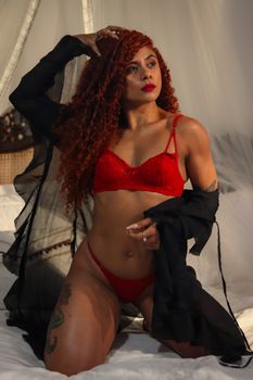 Confident woman posing in red lingerie and black robe on a bed indoors.