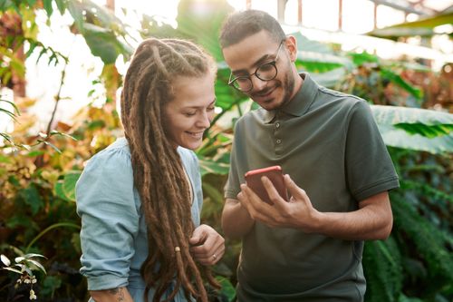 Two young adults smiling as they look at a phone together outdoors among green plants