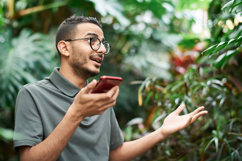 Landscaping contractor in gray polo shirt holding phone in voice recording position and gesturing toward lush garden plants while describing job scope