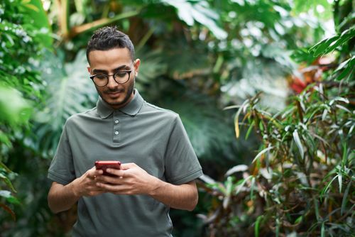 Free A young man wearing glasses uses a smartphone surrounded by vibrant green plants outdoors. Stock Photo