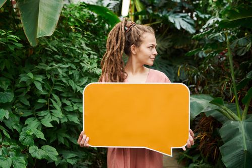 Free A woman with dreadlocks holds a blank orange sign in a vibrant green garden, providing perfect copy space. Stock Photo