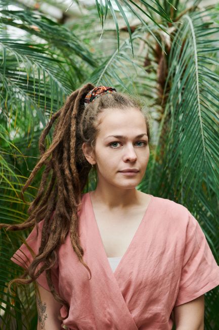 A woman with dreadlocks poses outdoors among lush plants, wearing a pink outfit.