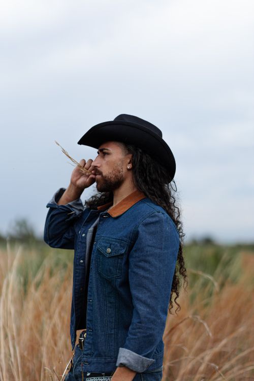 Free Portrait of a man wearing a cowboy hat and denim jacket standing in a grassy field. Stock Photo