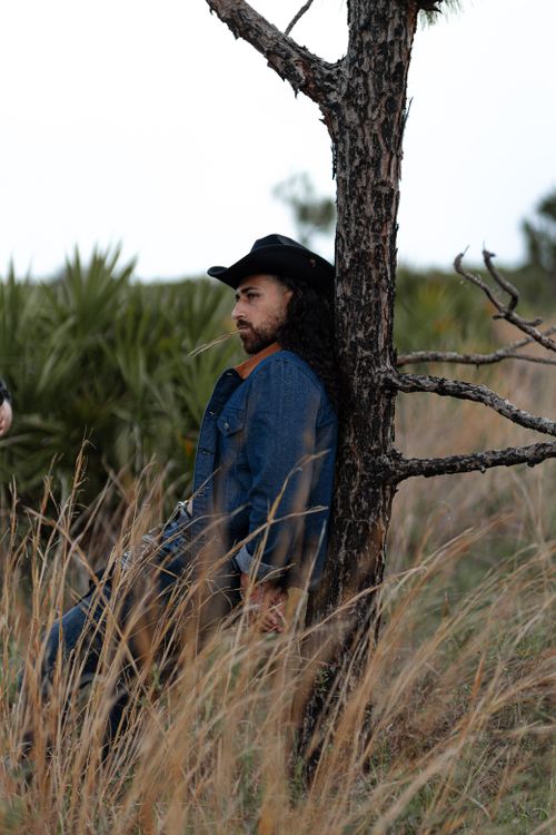 Free Man in denim and cowboy hat leaning against a tree in a grassy field. Stock Photo
