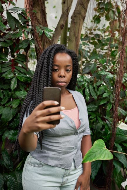 Young woman with braided hair taking a selfie amidst lush indoor plants.