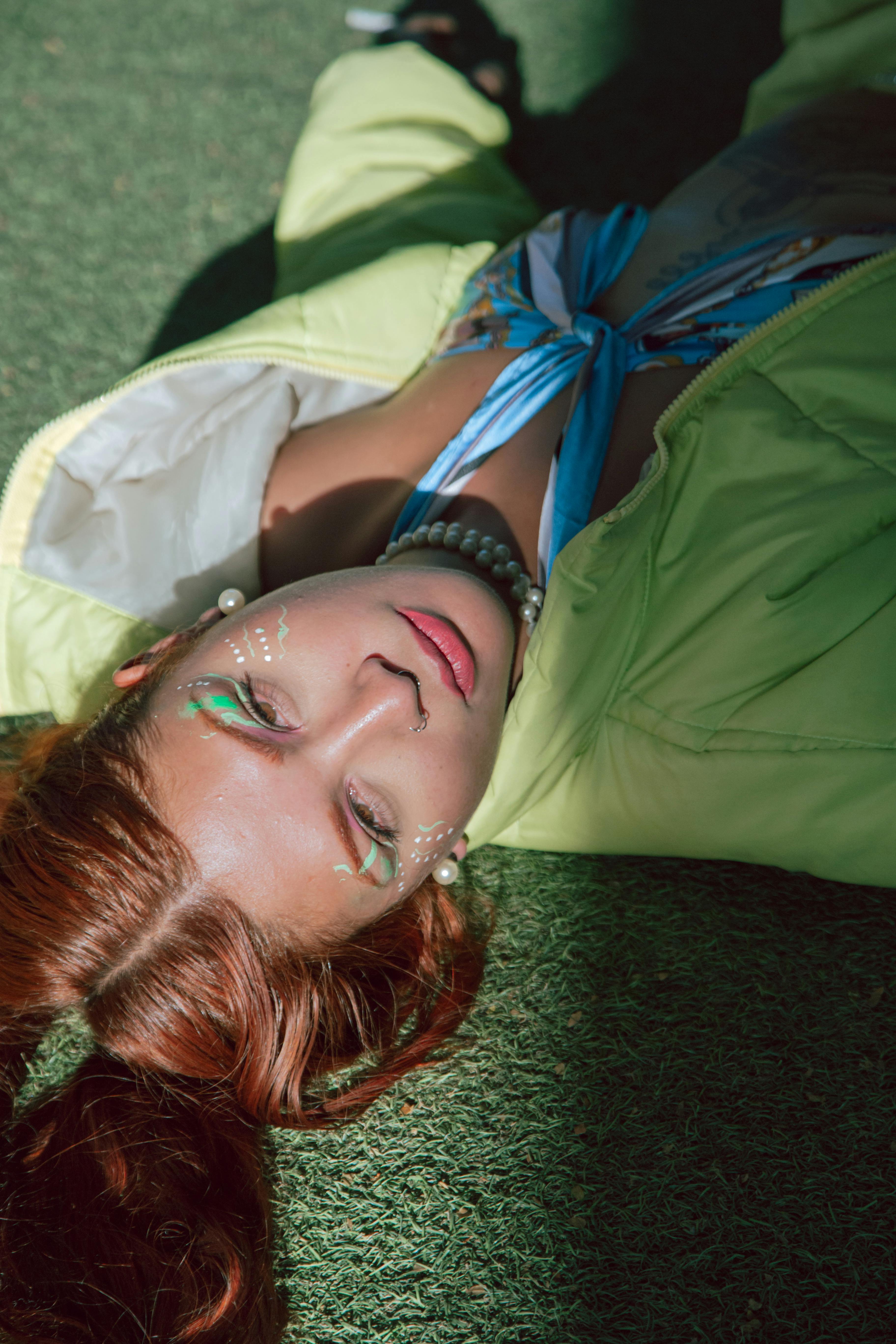 A young woman lies on grass in vibrant artistic makeup with natural light shadows.