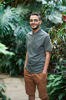 Smiling man in polo shirt and shorts standing in a vibrant garden surrounded by greenery.