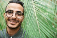 特写肖像中，一位戴眼镜的男子面带微笑，部分被绿叶遮挡，传递出温暖的感觉