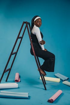 Stylish woman in black attire perched on a ladder against a blue studio background.