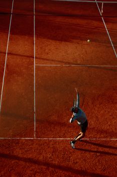 Aerial view of a tennis player serving on a clay court during a match at dusk.