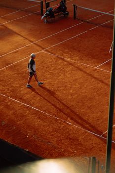 Adult male playing tennis on a clay court, captured at sunset, casting long shadows.
