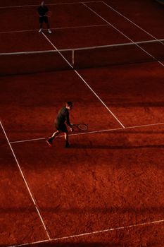 Two players compete on a clay tennis court during sunset, casting long shadows.