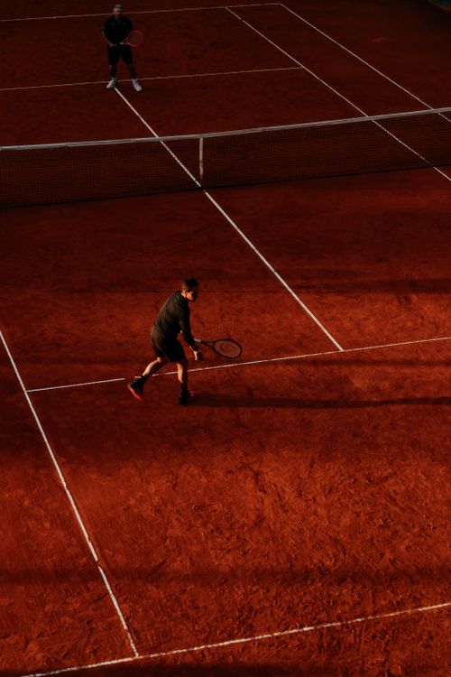Free Two players compete on a clay tennis court during sunset, casting long shadows. Stock Photo