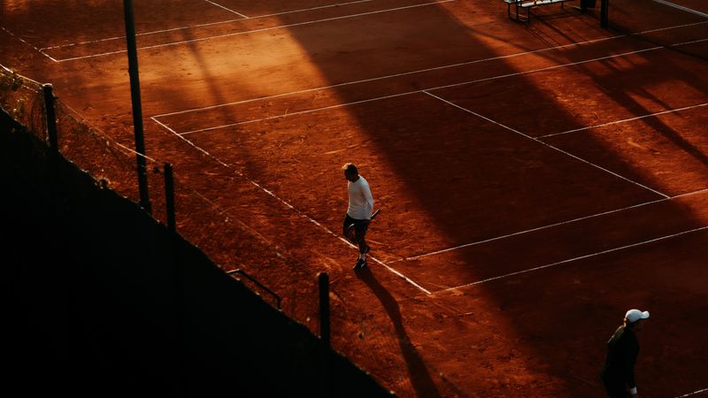 Dynamic aerial shot of a tennis match on a clay court during sunset, capturing shadows and movement.