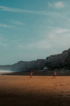 Two individuals enjoy a game of ball on a scenic beach with cliffs and a serene ocean backdrop.