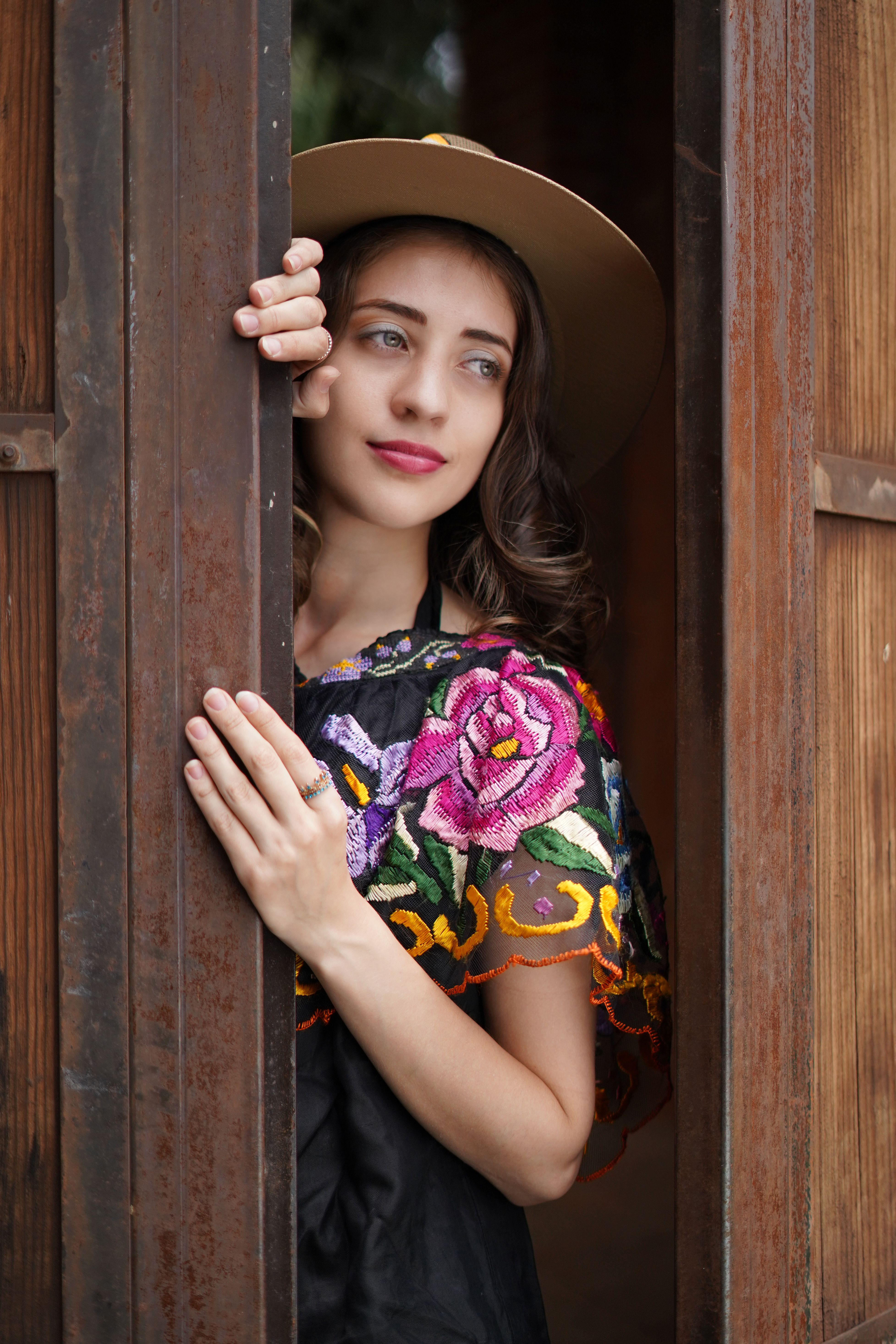 Young woman in floral embroidered top peeking through a wooden doorway, wearing a hat.