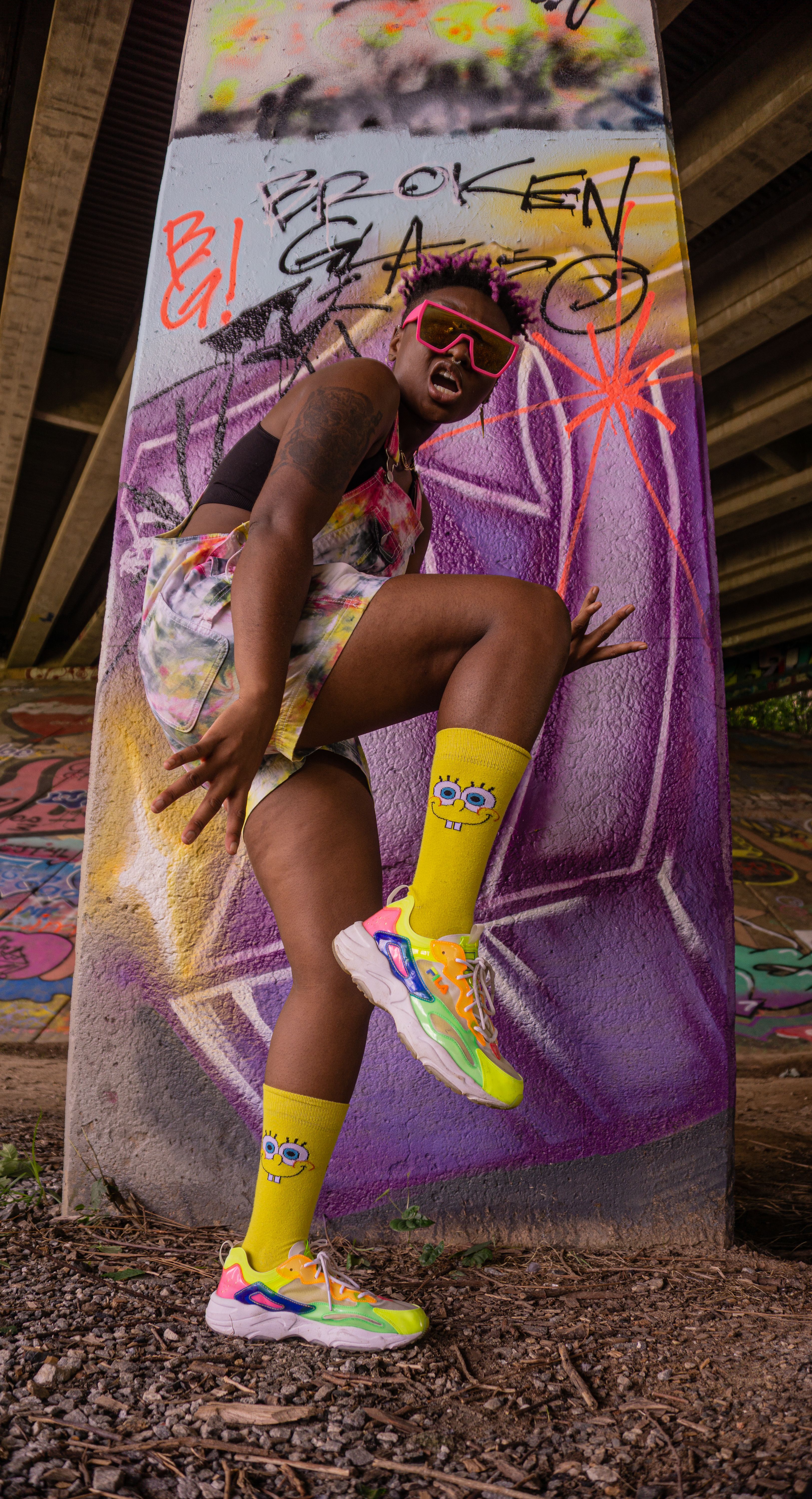 A joyful skater poses energetically in colorful attire against a graffiti-covered pillar.