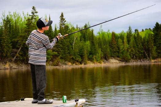 Young boy fishing with a rod on a wooden dock surrounded by lush greenery in Dinorwic, Ontario.