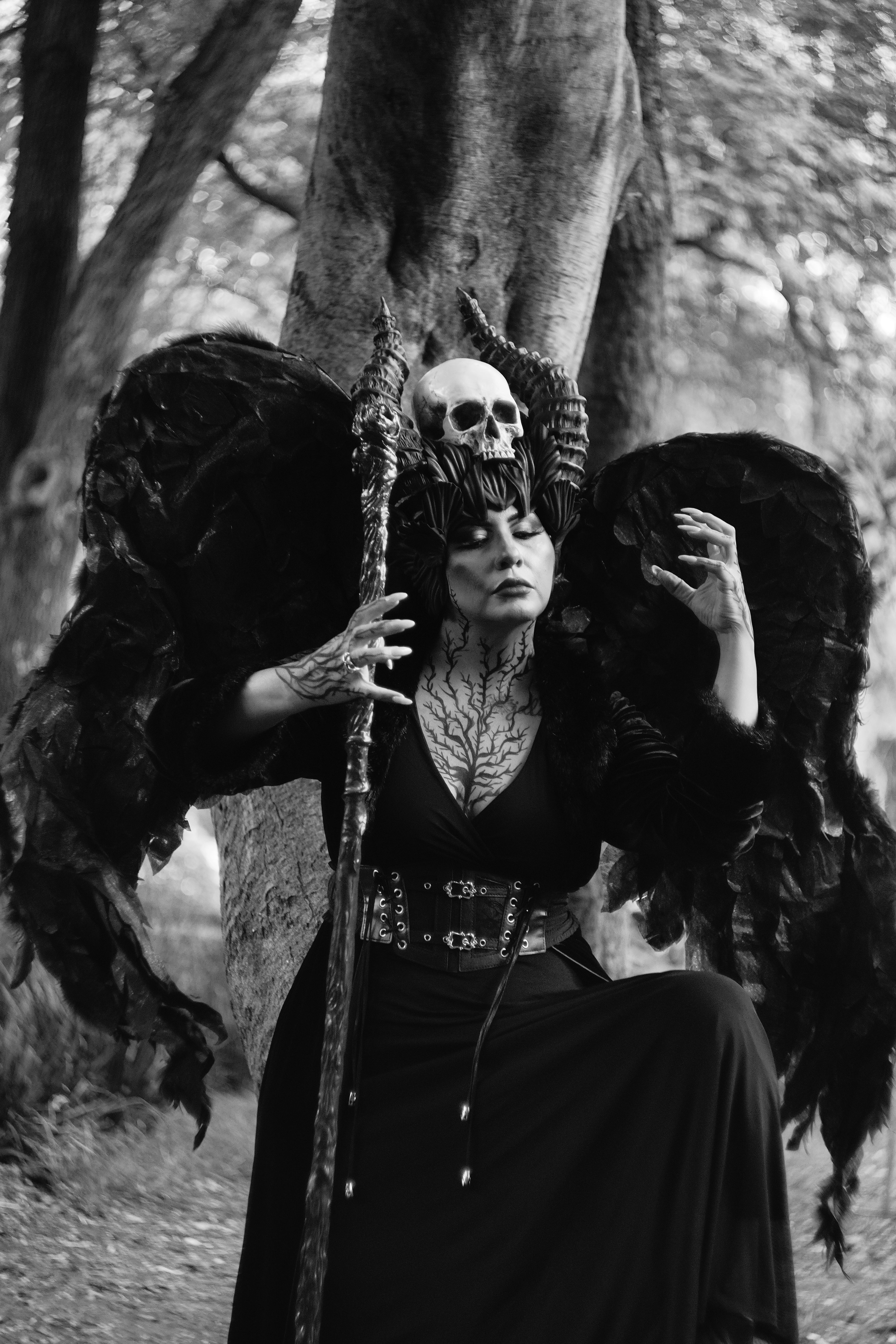 Gothic woman with skull headdress posing in a forest setting, black and white.