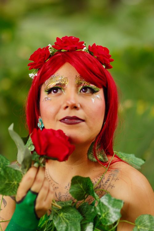Free A woman with floral headdress and glittering makeup clutching a red rose amidst lush greenery. Stock Photo
