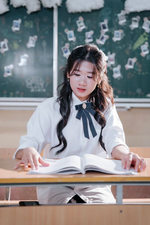 Free Young woman in uniform focused on reading at school desk with a decorated chalkboard background. Stock Photo