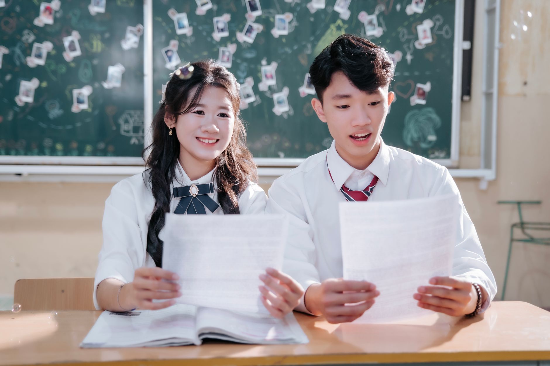 A smiling boy and girl in uniform reading papers in a classroom setting.