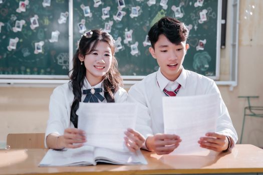 A smiling boy and girl in uniform reading papers in a classroom setting.