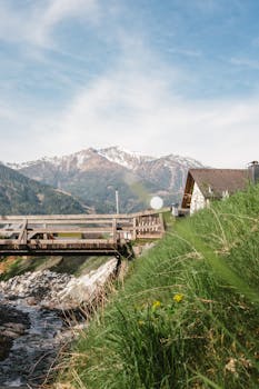 Tranquil Alpine scene with a cottage, bridge, and snowy mountains.