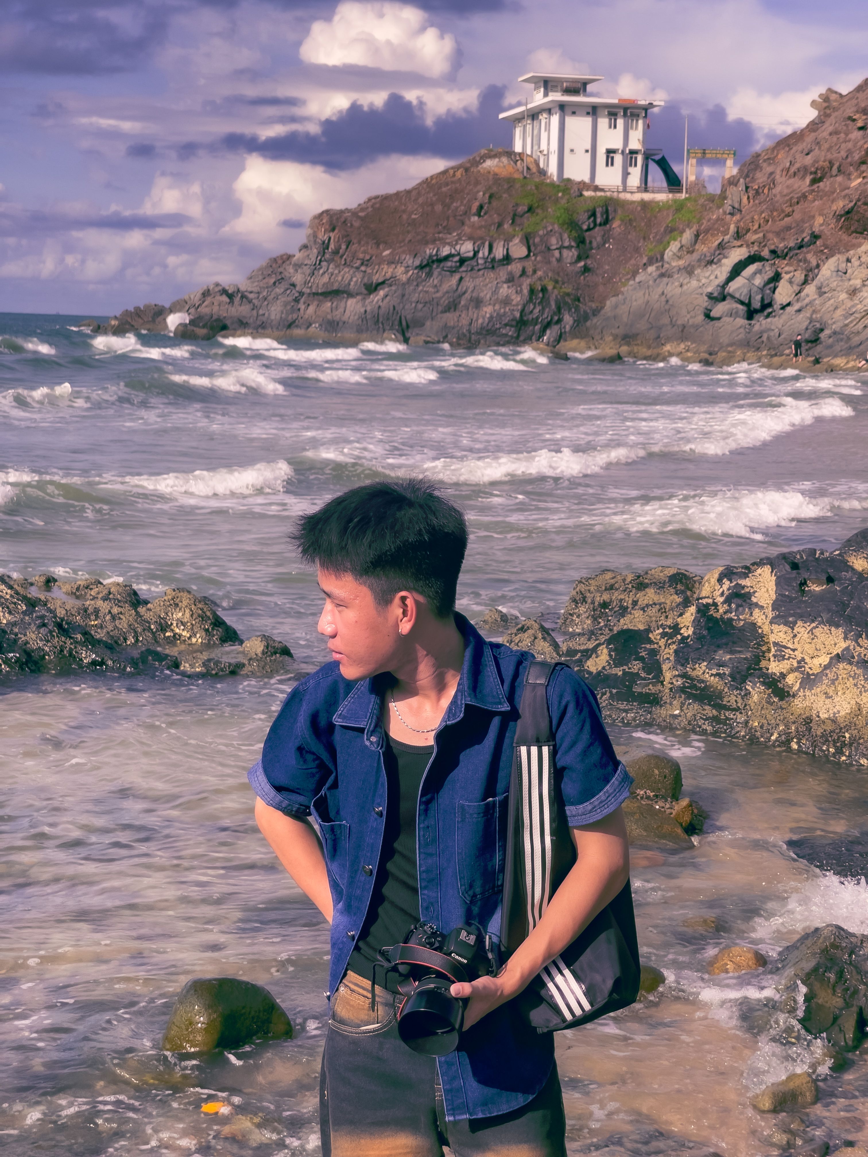 Young man with camera on a rocky shore near a modern coastal house, under a cloudy sky.