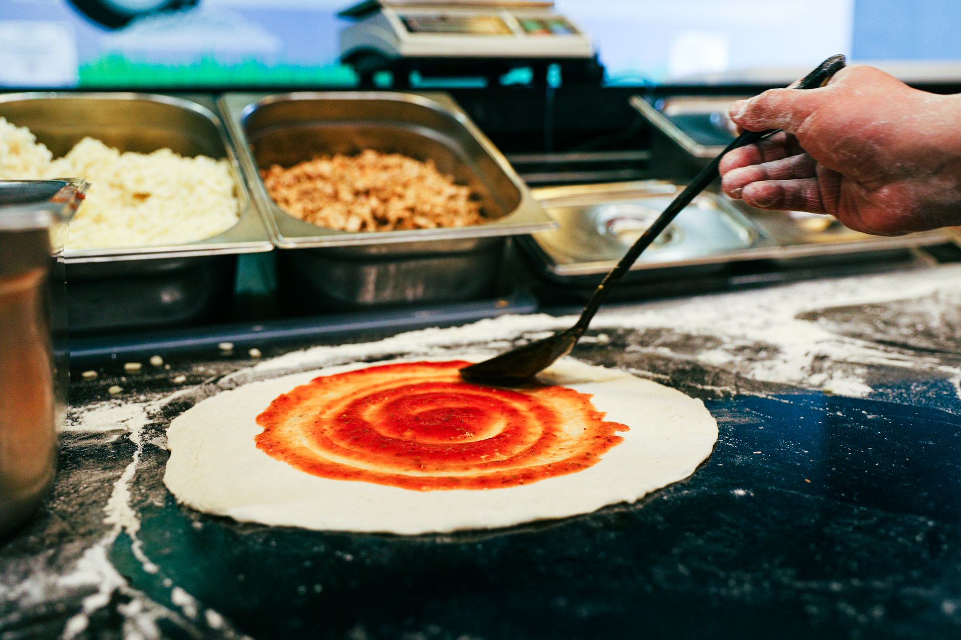 Chef spreading tomato sauce on pizza dough in kitchen setting.
