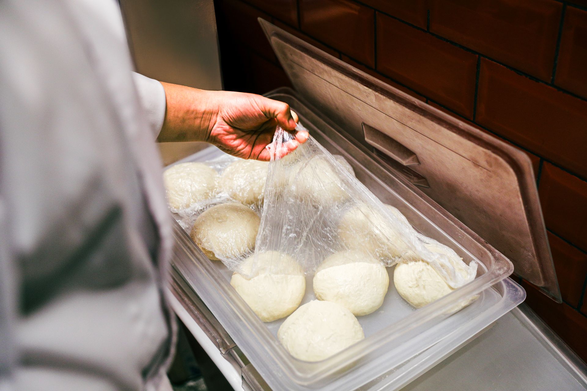 Chef lifting plastic wrap from fresh dough balls in a kitchen setting.