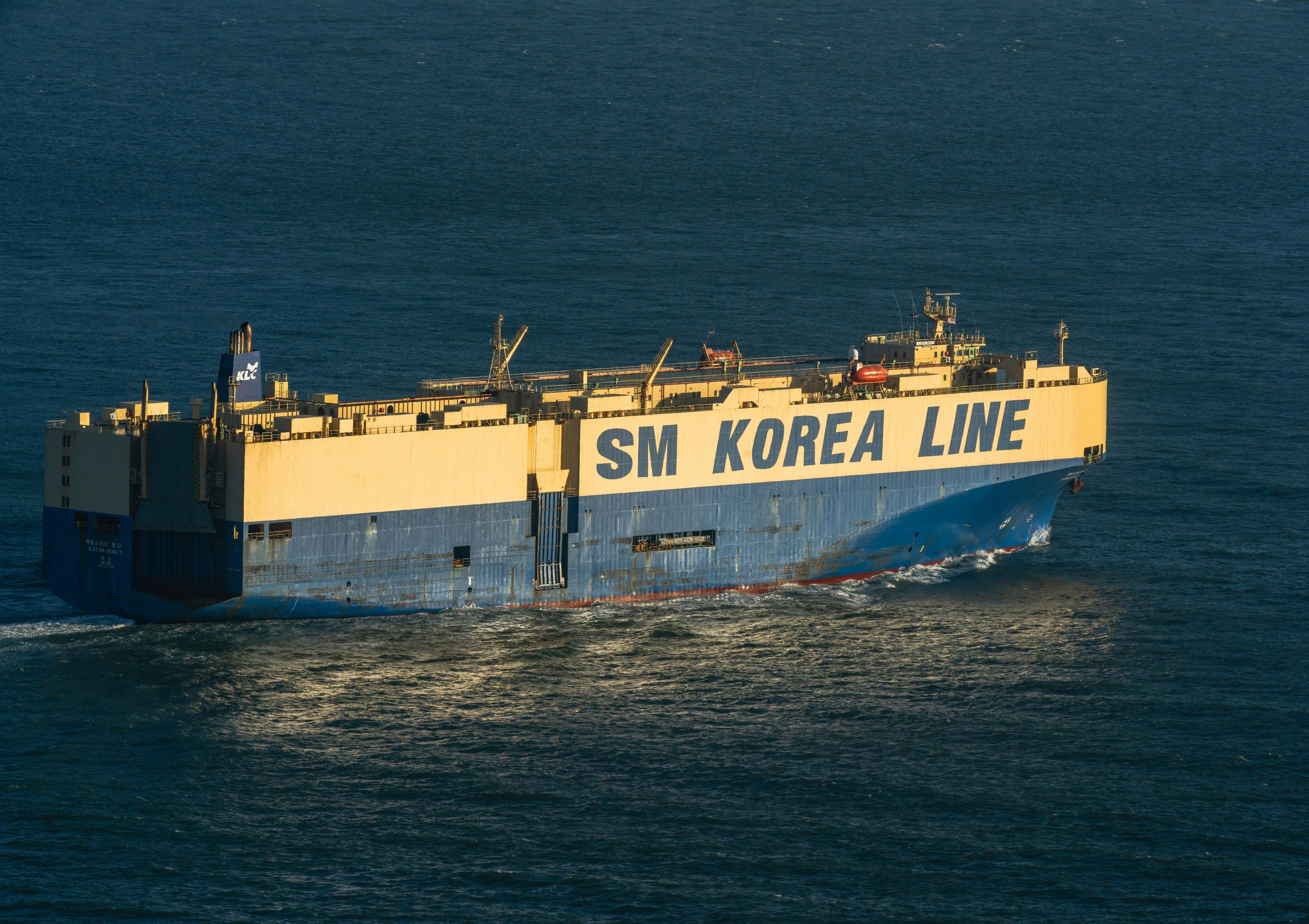 A large cargo ship, SM Korea Line, navigating through calm ocean waters under a clear blue sky.