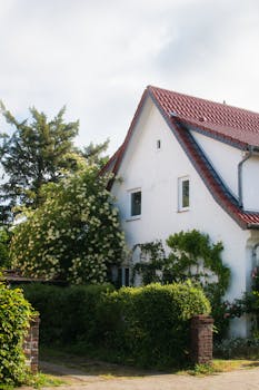 Beautiful white cottage with red roof surrounded by lush greenery and blooming shrubs on a sunny day.
