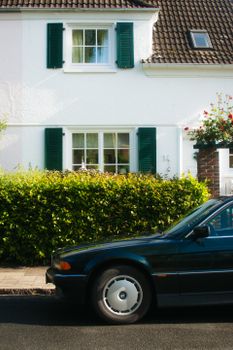 White suburban house with green shutters and classic car parked outside on a sunny day.