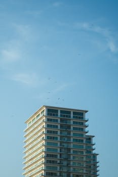 A contemporary high-rise building against a bright blue sky with clouds and birds.