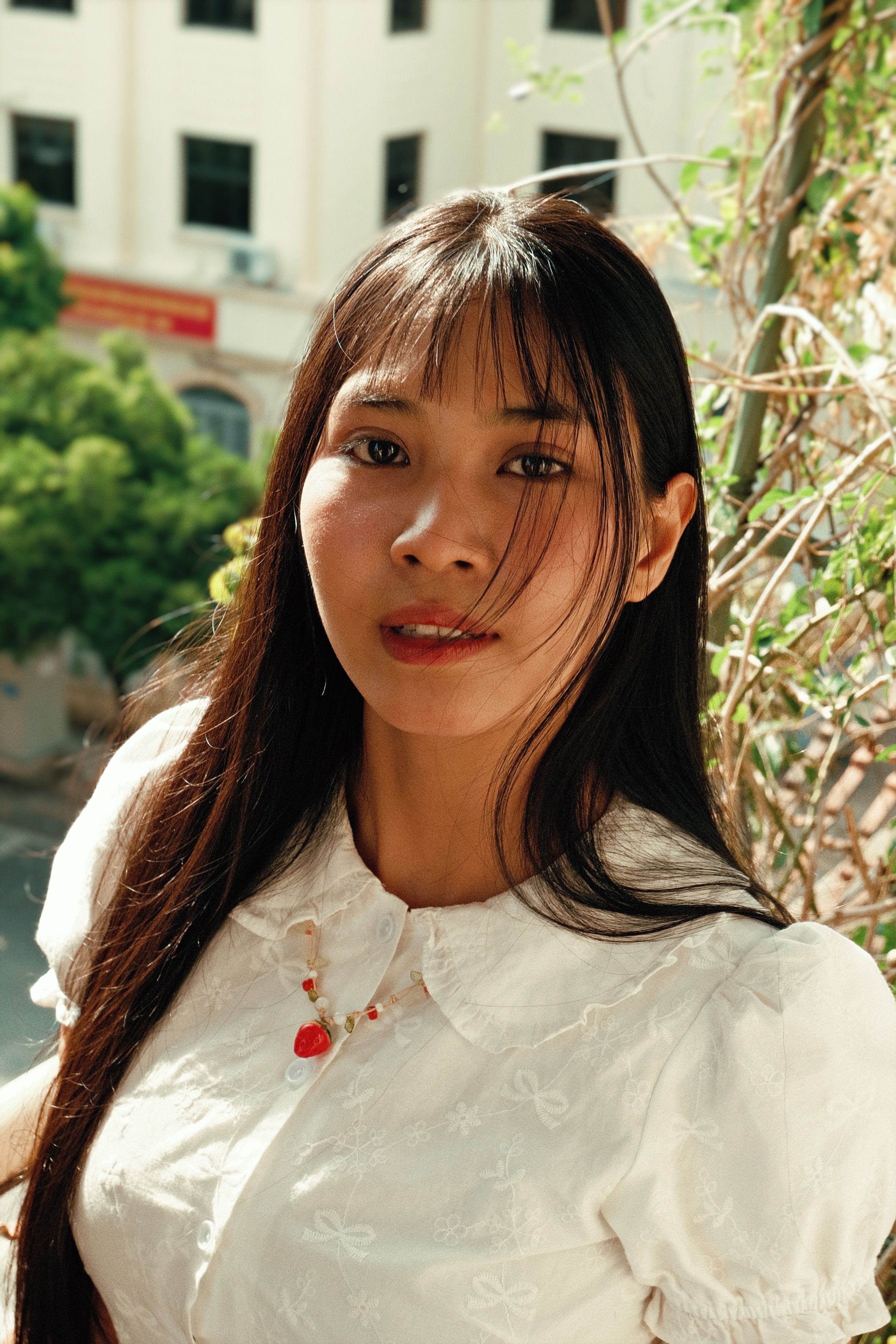 A young woman in a white blouse stands outdoors, looking at the camera.