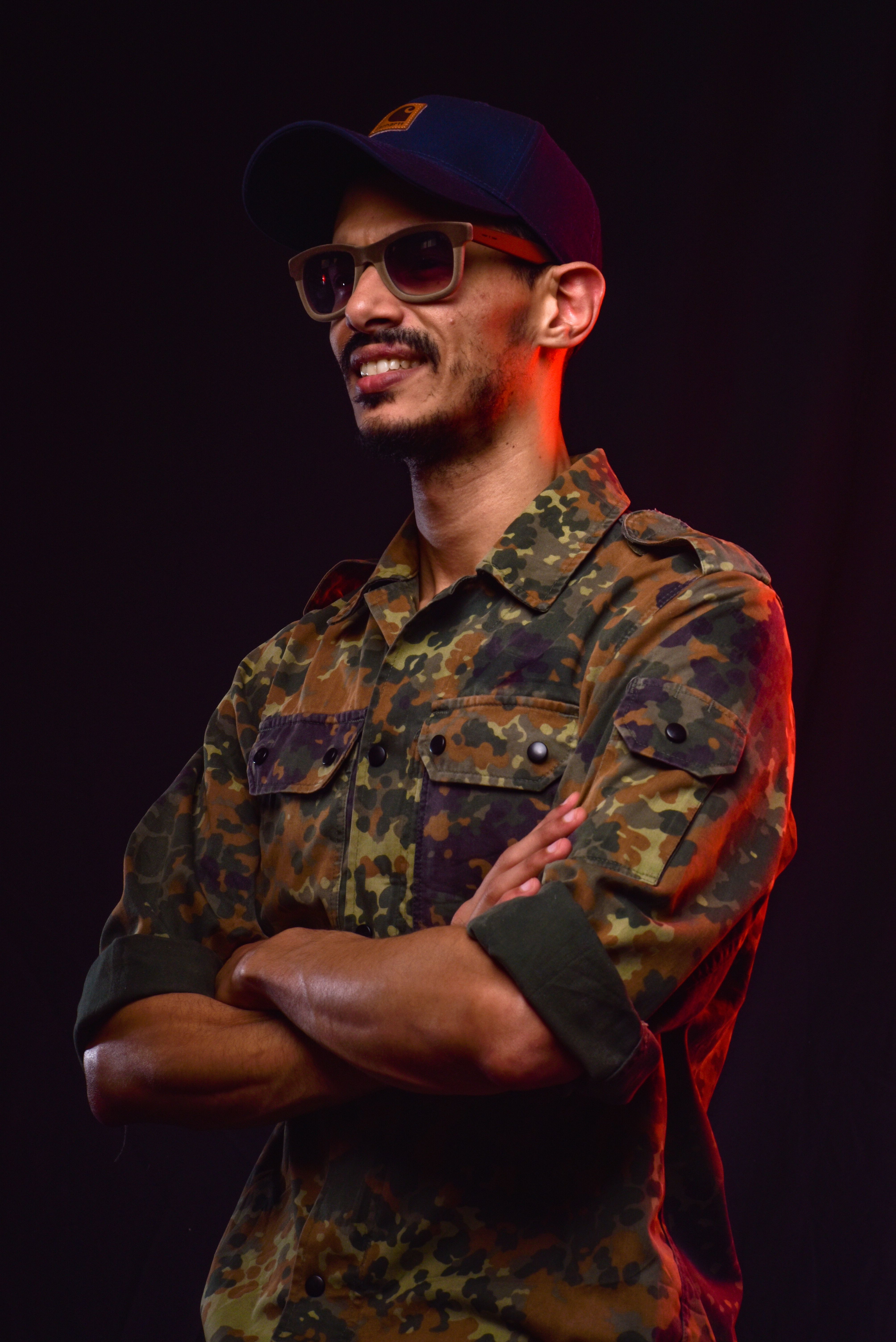 Portrait of a man in a camouflage shirt, arms crossed confidently under dramatic lighting.
