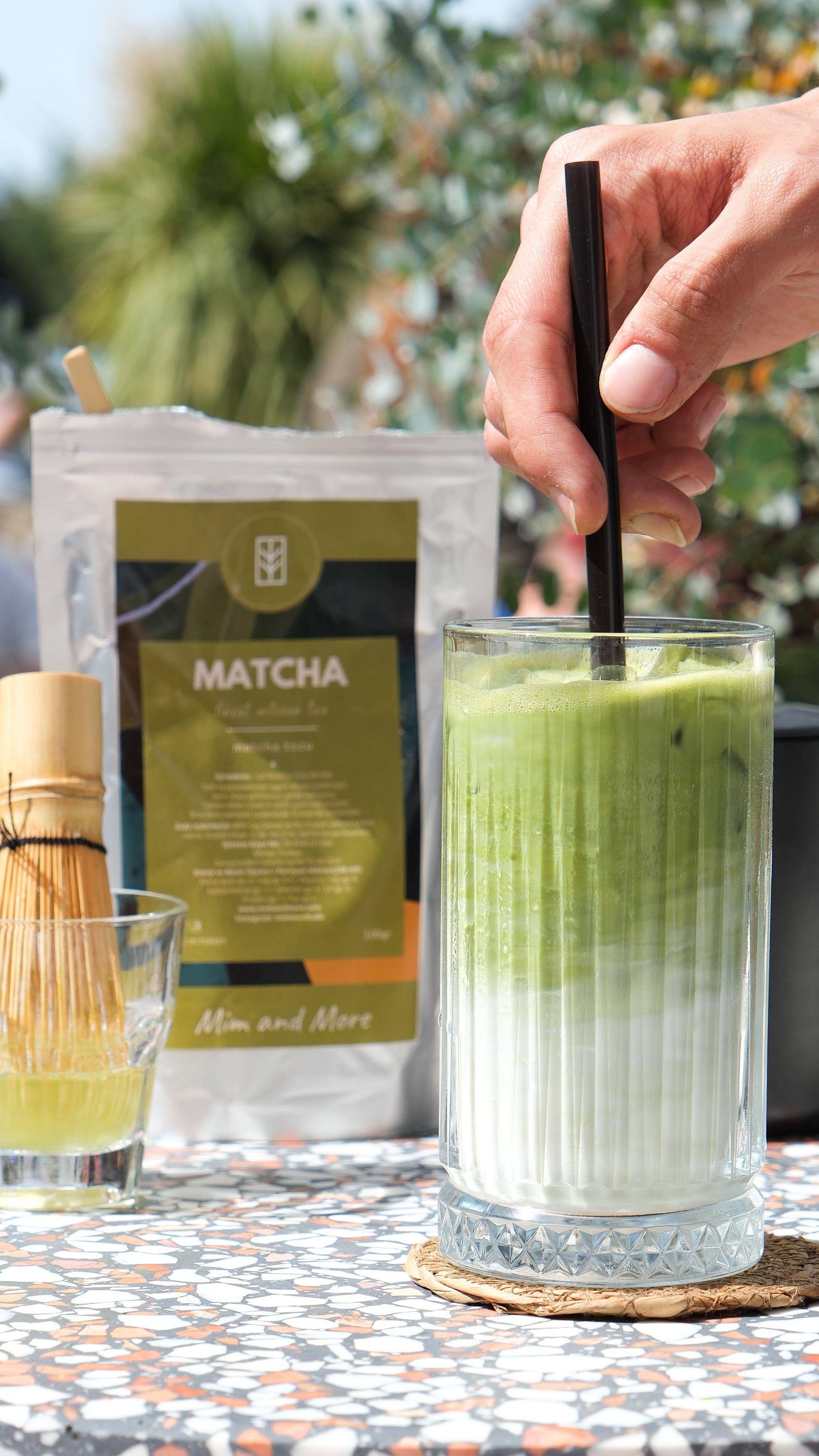 A hand stirs a refreshing iced matcha latte outdoors, highlighting the vibrant green and white layers.